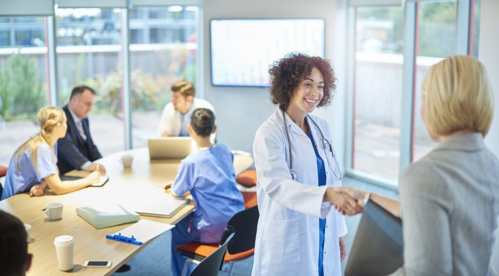 A doctor in a white coat shakes hands with a woman in an office, highlighting the importance of effective Training Solutions, while other medical professionals sit at a table in the background.