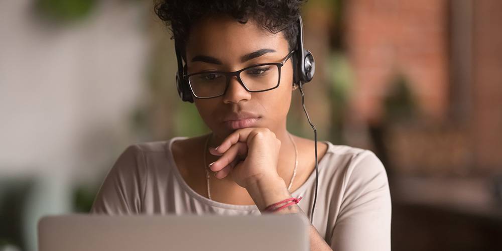 A person wearing glasses and headphones looks intently at a laptop screen, resting their chin on their hand.