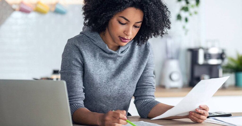A woman sits at a table, reviewing a document and taking notes with a pencil. A laptop and other papers are on the table in front of her.