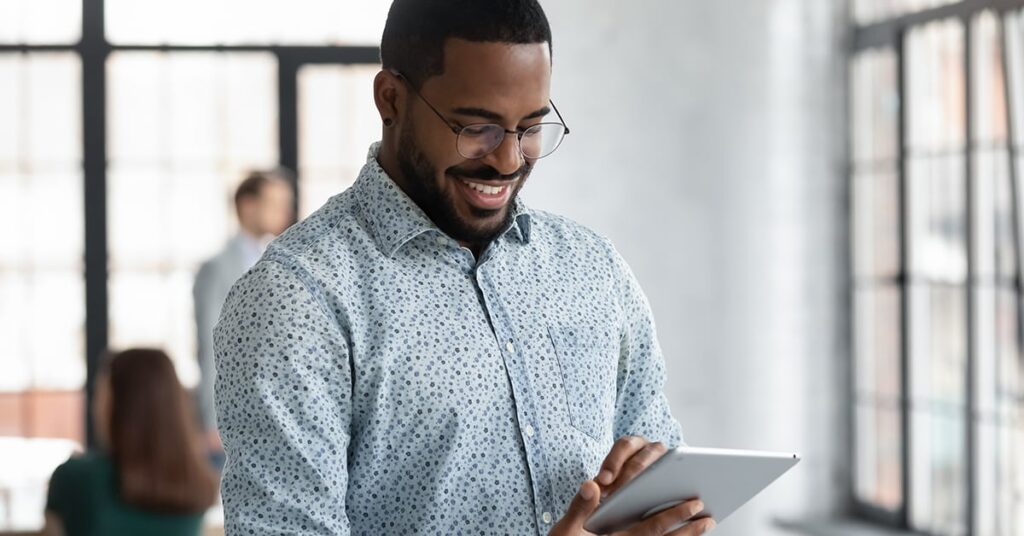 A man wearing glasses and a patterned shirt stands indoors, smiling while using a digital tablet. Blurred people and large windows are visible in the background.