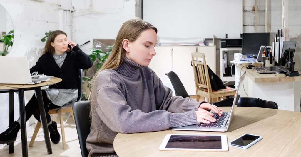 Two women working in a modern office; one types on a laptop at a round table with a tablet and phone, while the other talks on the phone at a nearby desk.