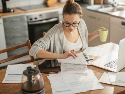 A person sits at a kitchen table reviewing papers, holding a mug, with a laptop and documents spread out in front of them.