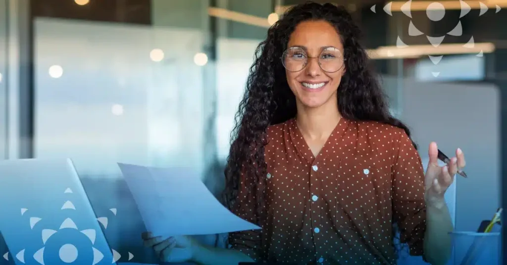 A woman with long curly hair and glasses, wearing a brown polka dot shirt, smiles while holding a sheet of paper in an office setting.