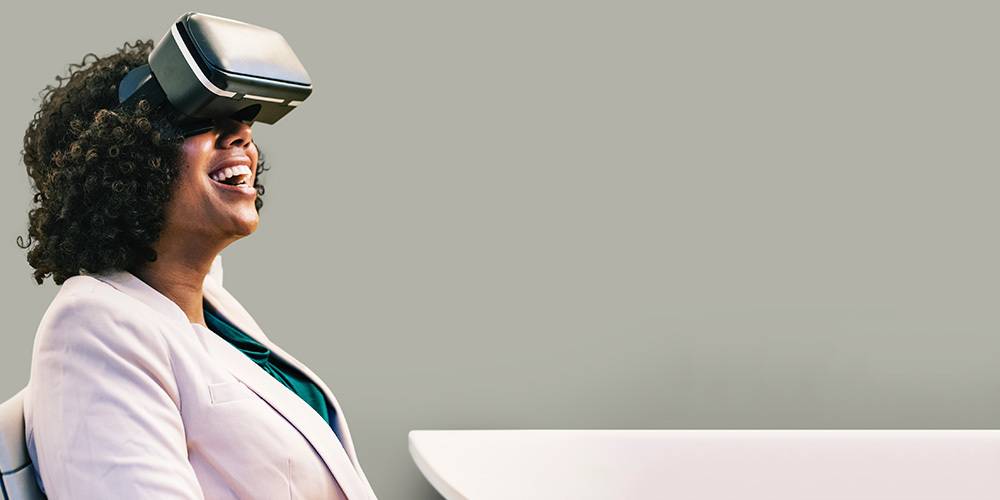 A woman sits at a desk, smiling and wearing a virtual reality headset, against a plain background.