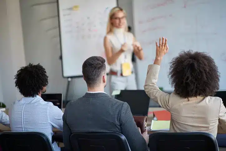 Three people sit facing a presenter at a whiteboard; one attendee raises their hand, indicating a question or participation in the discussion.