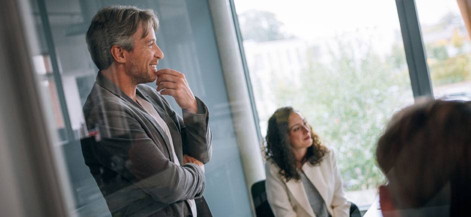 A man stands and smiles while a woman sits and listens during a meeting in a modern office with large windows.