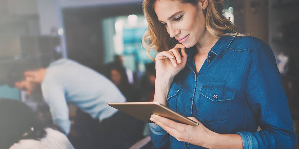 A woman in a denim shirt stands indoors, looking at a tablet. Other people are working in the background.