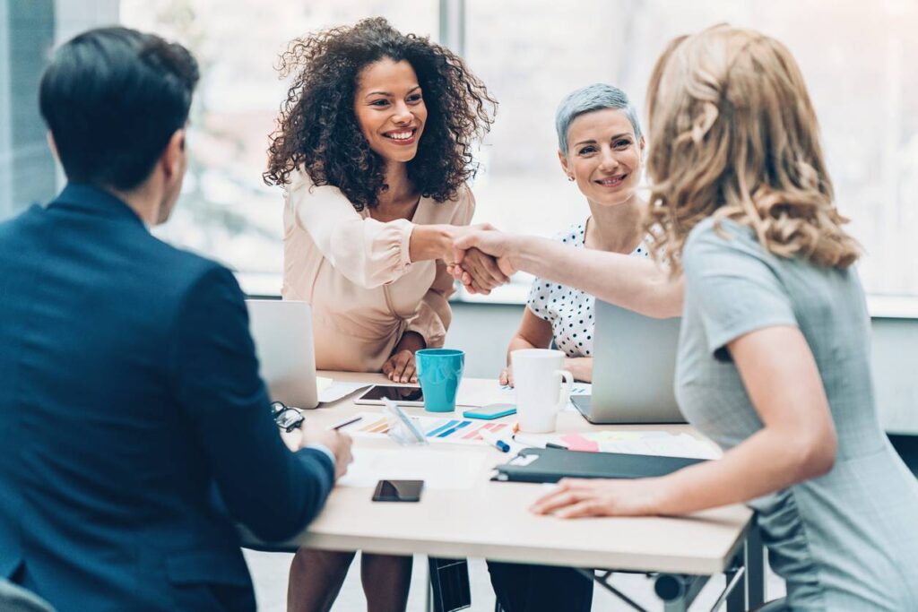 Four people in business attire sit at a table with laptops and papers; two women in the center shake hands and smile at each other.