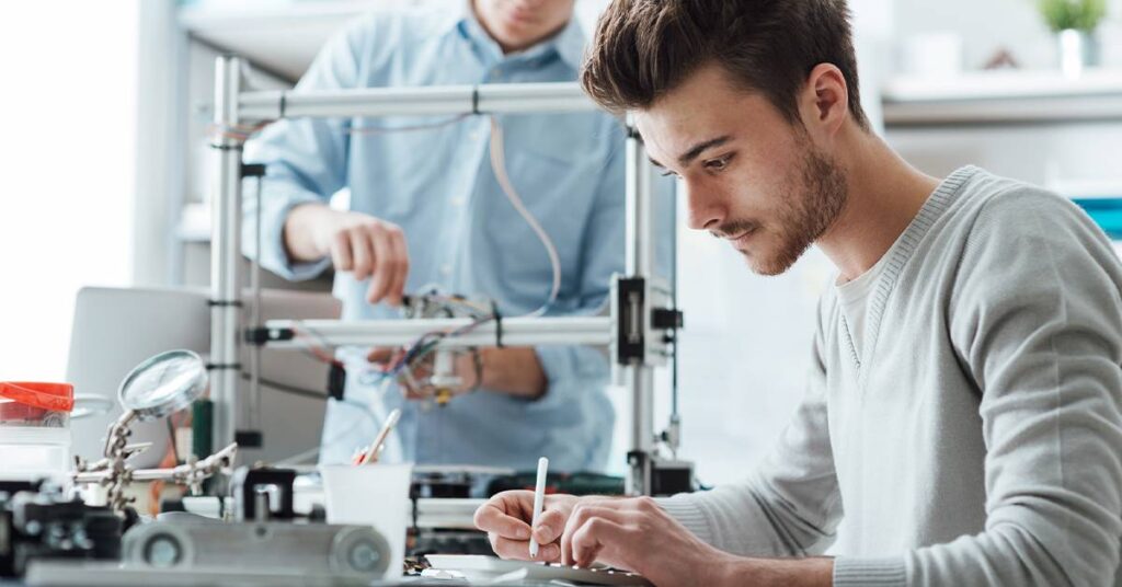 Two men work in a lab; one writes on a notepad while the other adjusts a piece of equipment, possibly a 3D printer, in the background.