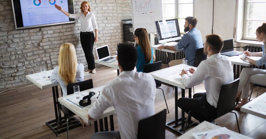 A woman presents data on a screen to a group of people seated at desks with laptops in a modern classroom setting.