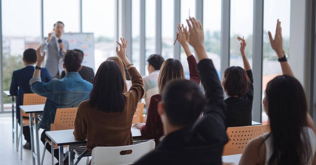 Students seated in a classroom raise their hands while a teacher stands at the front, speaking and gesturing near large windows.