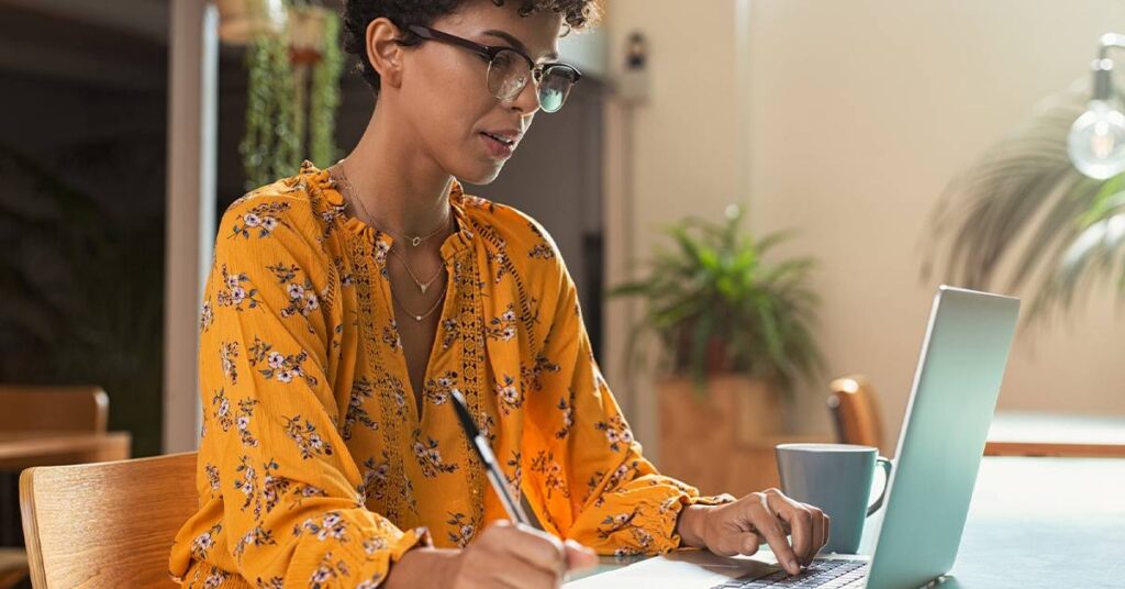 A woman in a yellow floral blouse works at a laptop, writing in a notebook with a pen. A mug and indoor plants are visible in the background.
