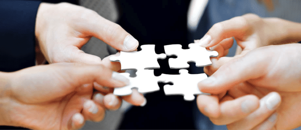 Four hands each hold a puzzle piece, joining them together to complete a small section of a puzzle.