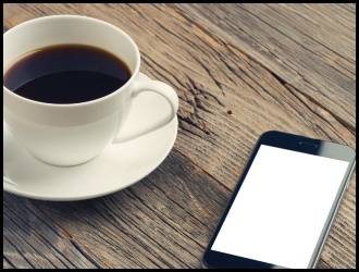 A cup of black coffee in a white mug sits on a wooden table next to a smartphone with a blank white screen.