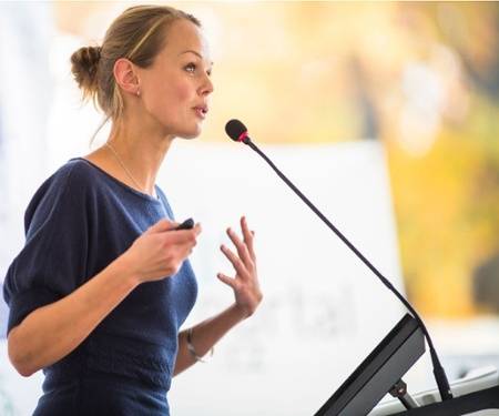 Woman speaking at a podium with a microphone, holding a small device, gesturing with one hand, and looking attentive.