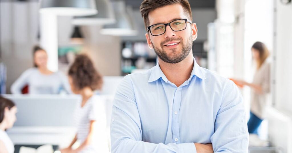 Man with glasses and beard in a light blue shirt stands with arms crossed in an office, with several people working in the background.