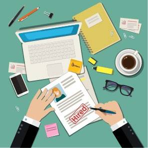 A person signs a job offer letter stamped "Hired" at a desk with a laptop, coffee, notebook, glasses, phone, and office supplies.