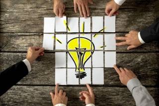 Five hands arrange square cards on a wooden surface to form a drawing of a lit yellow light bulb.