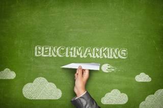 A hand holding a paper airplane is pointed toward the word "BENCHMARKING" on a green chalkboard background with cloud drawings.