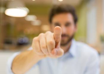 A man in a blurred background points his index finger directly at the camera, with his face out of focus.