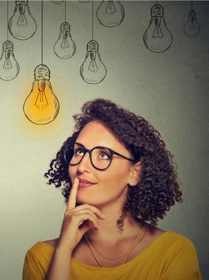 Woman with curly hair and glasses looks thoughtful with finger on chin, standing in front of drawn light bulbs, one of which is illuminated.