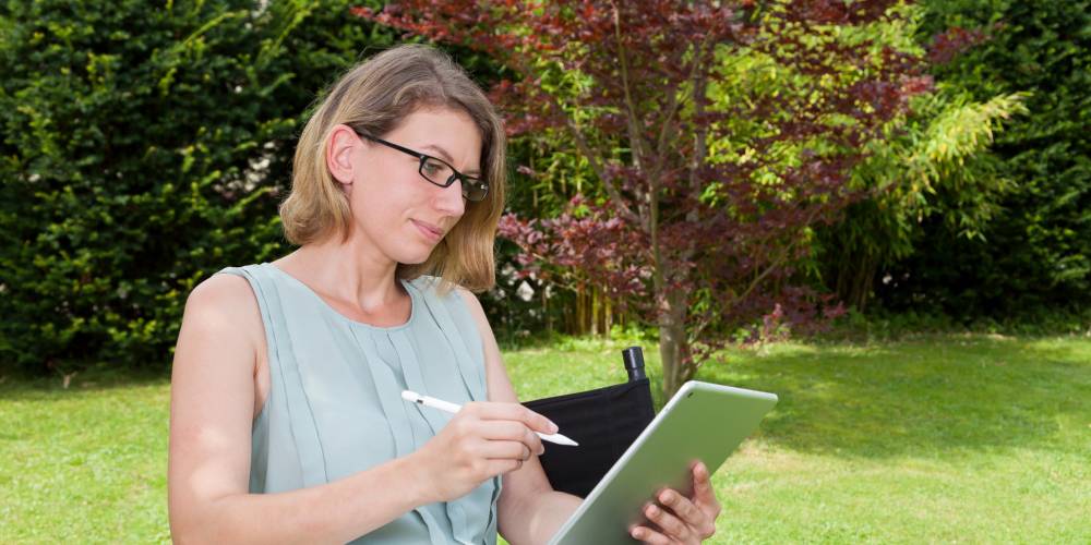A woman sits outdoors on a chair, using a tablet with a stylus in a garden setting with trees and grass in the background.
