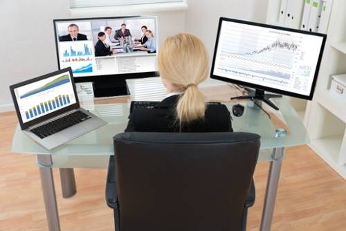 A person sits at a desk with a laptop and two monitors displaying charts, graphs, and a video conference with four people.
