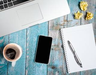 A laptop, smartphone, notepad with pen, cup of coffee, and two crumpled yellow papers on a blue wooden surface.