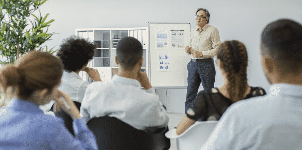 A person stands and presents charts on a flipchart to a seated group in a modern office setting.
