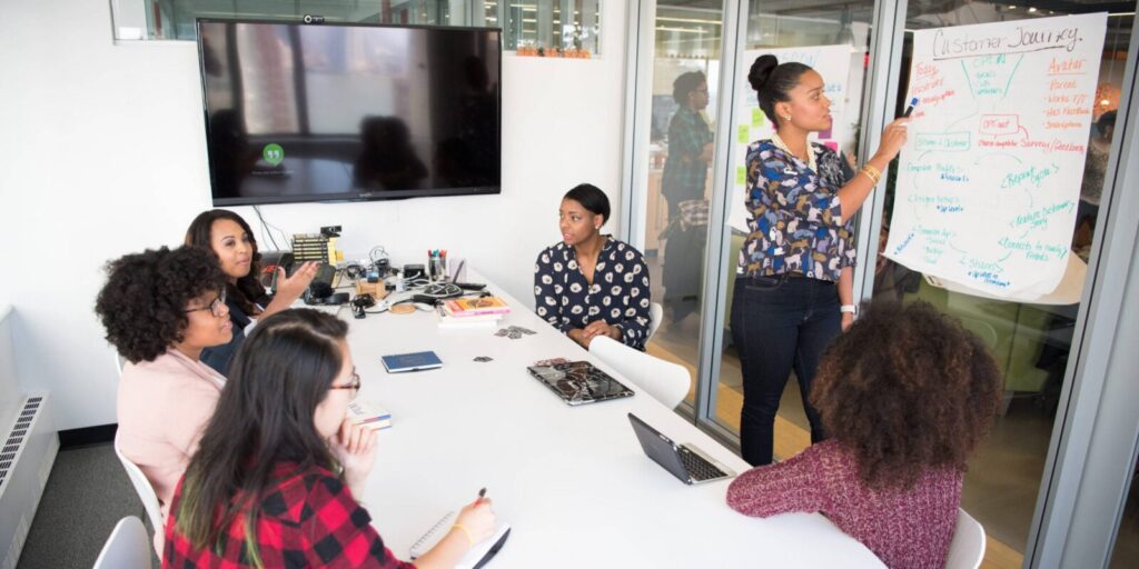 A woman stands and writes on a flip chart while five colleagues sit around a conference table watching and listening during a meeting in a modern office.