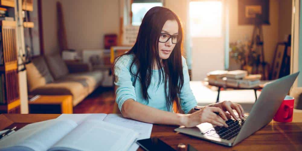 A woman with long dark hair and glasses types on a laptop at a desk with papers, a notebook, and a red mug in a sunlit home office.