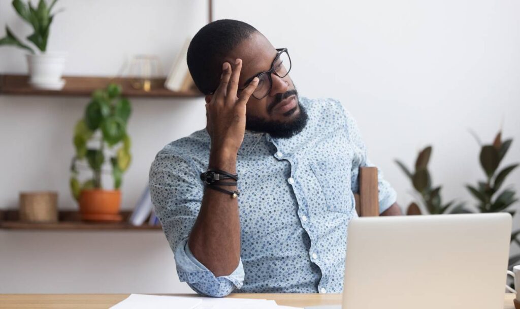 A man wearing glasses sits at a desk with a laptop, resting his head on his hand and appearing thoughtful or stressed. Indoor plants and shelves are in the background.