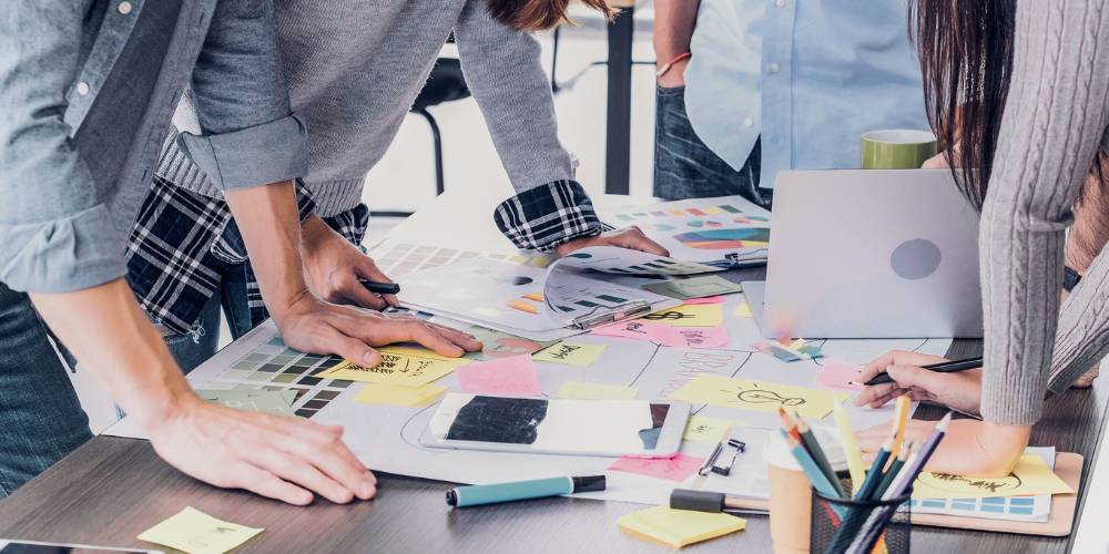 Several people gather around a table covered with documents, color samples, sticky notes, a smartphone, a laptop, and stationery, collaborating on a project.