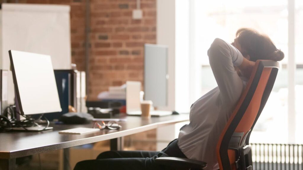 Person sitting at an office desk, leaning back in their chair with hands behind their head, looking relaxed. Computer monitor and office items visible on the desk.