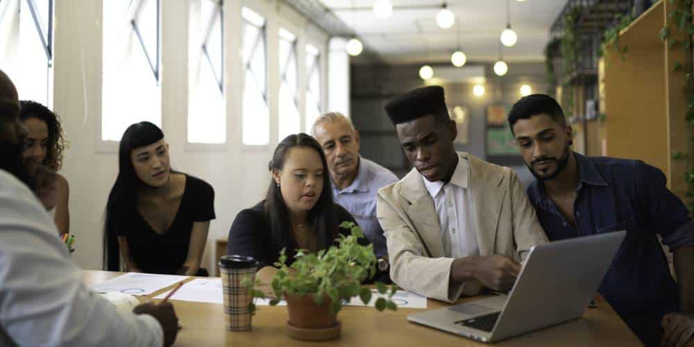 A group of six people sits at a table, looking at a laptop screen together in a well-lit office space.