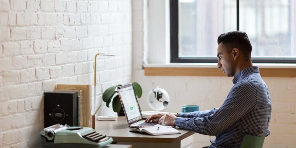 A man sits at a desk working on a laptop in a modern office with a brick wall, window, a globe, books, a plant, and a typewriter.