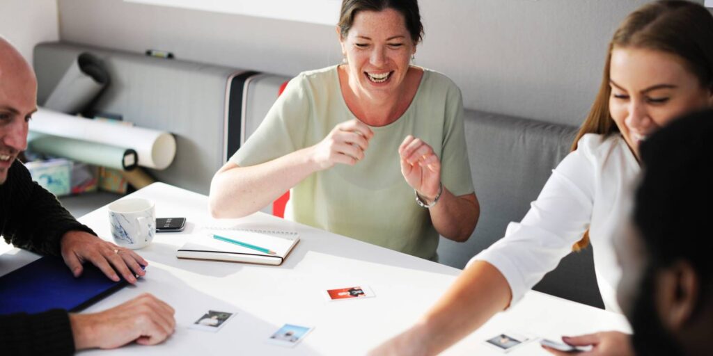 Four people sit around a white table, smiling and engaging with photos and papers, suggesting a collaborative meeting or group activity.