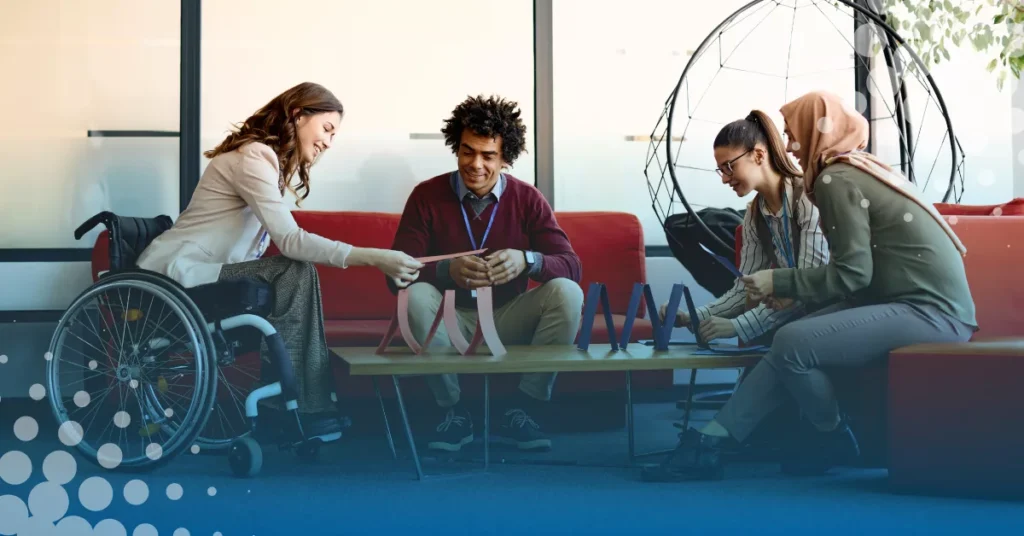 Four people, including a woman in a wheelchair, collaborate around a low table with paper structures in a modern office lounge area.