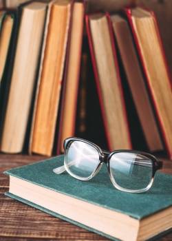 A pair of eyeglasses rests on a closed book with more books standing upright in the background.