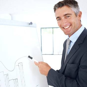 Man in a suit smiling and pointing at a chart with a marker on a flipchart in a bright office setting.