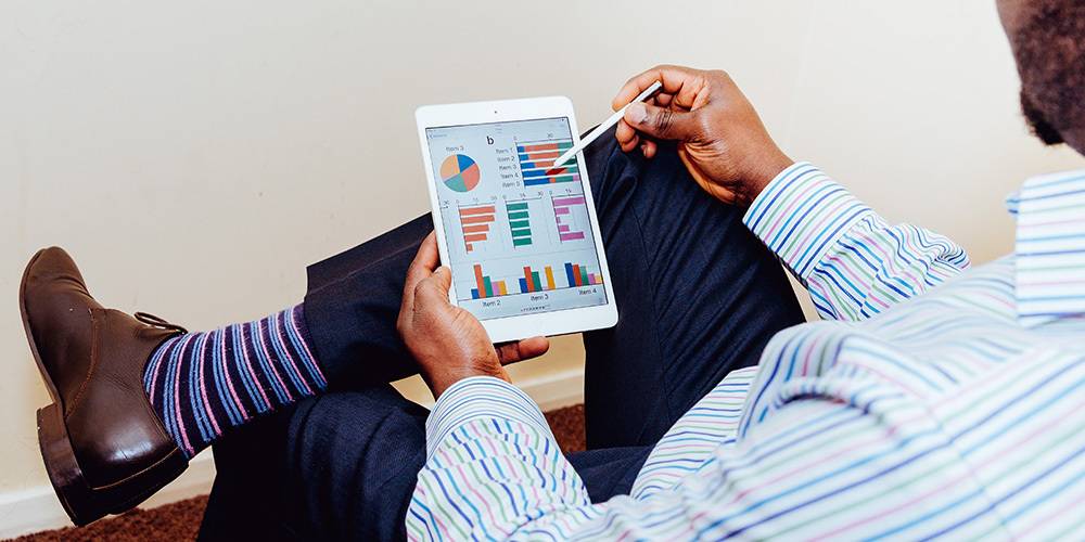A person in business attire reviews colorful charts and graphs on a tablet using a stylus, sitting with legs crossed.