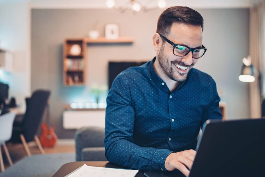 A man wearing glasses and a blue shirt is smiling while working on a laptop at a desk in a modern, well-lit living room.