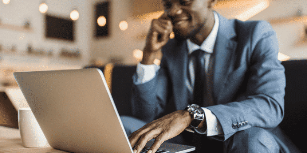 A man in a suit sits at a table, smiling while using a laptop. A white cup is on the table, and the background is softly lit.