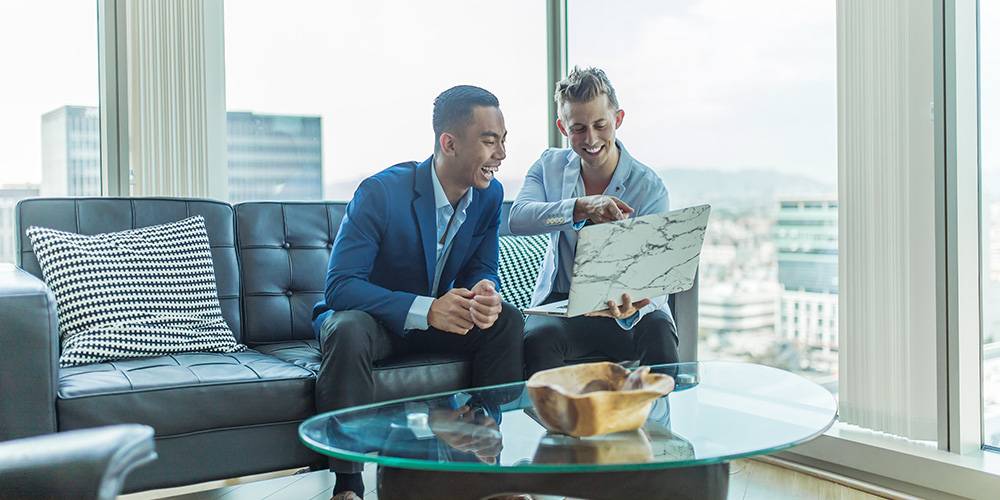 Two men in business attire sit on a sofa in a modern office, looking at a laptop and smiling, with large windows and city buildings in the background.