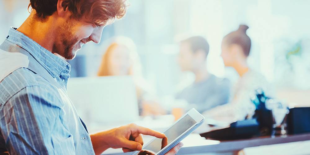 Man using a tablet and smiling in the foreground, with three people having a discussion at a table in a blurred office background.