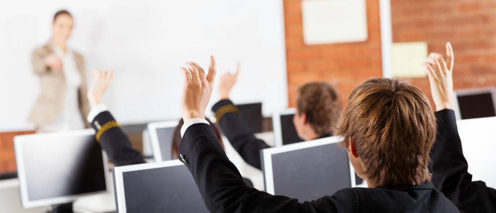 Students in a classroom raise their hands while seated at desks with computers, facing a teacher standing at the whiteboard.