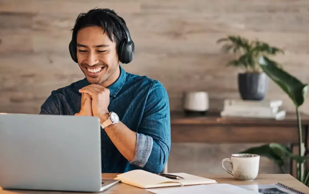 A person wearing headphones sits at a desk with a laptop, notebook, and coffee mug, smiling while looking at the screen in a home office setting.