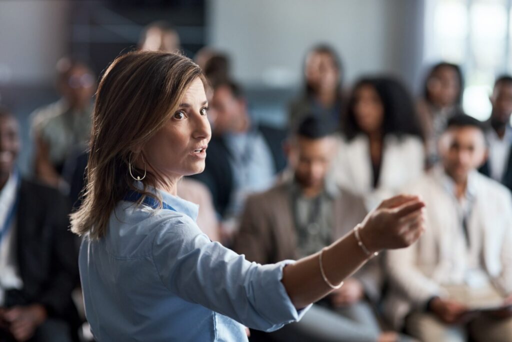 A woman in business attire gestures while speaking to a seated audience in a conference or meeting setting.