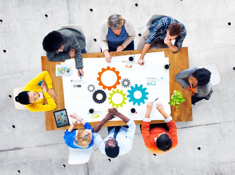 Eight people sit around a table with colorful gear diagrams, discussing and collaborating on a project, viewed from above.