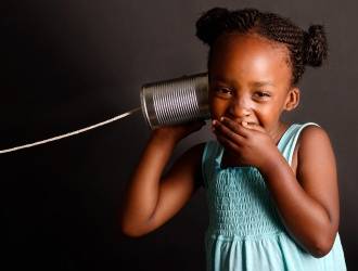 Young girl in a blue dress smiles and covers her mouth while holding a tin can phone to her ear, against a dark background.